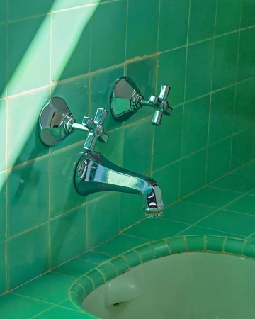 Close-up of a chrome faucet with cross handles mounted on a green tiled wall and sink edge, with bright light casting shadows.