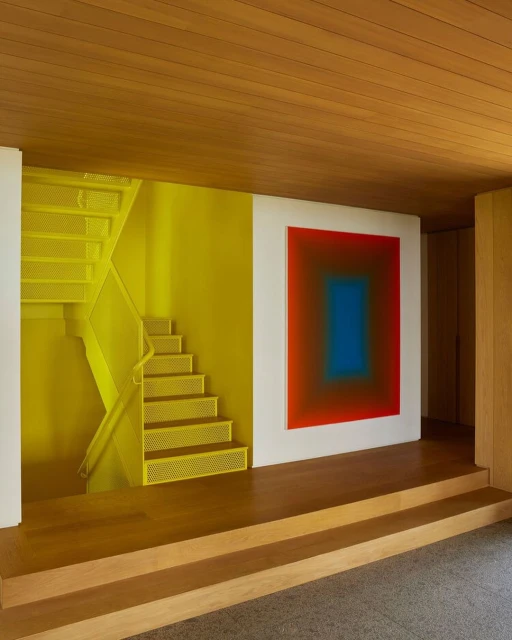 Indoor shot of yellow metal staircase on a yellow wall adjacent to an abstract square painting in a modern home interior.