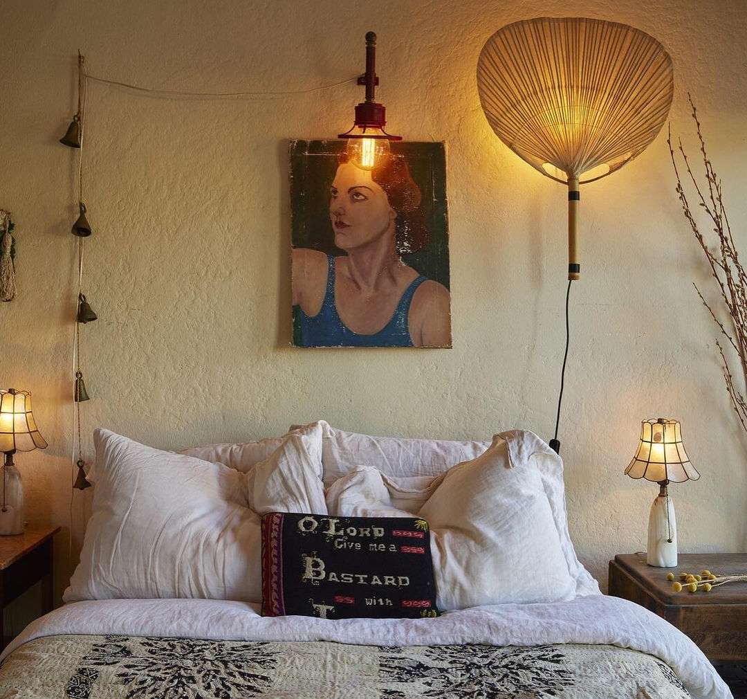 Bedroom interior with bed, pillows, lamps, and artwork featuring a portrait and decorative hanging elements in a cozy setting.