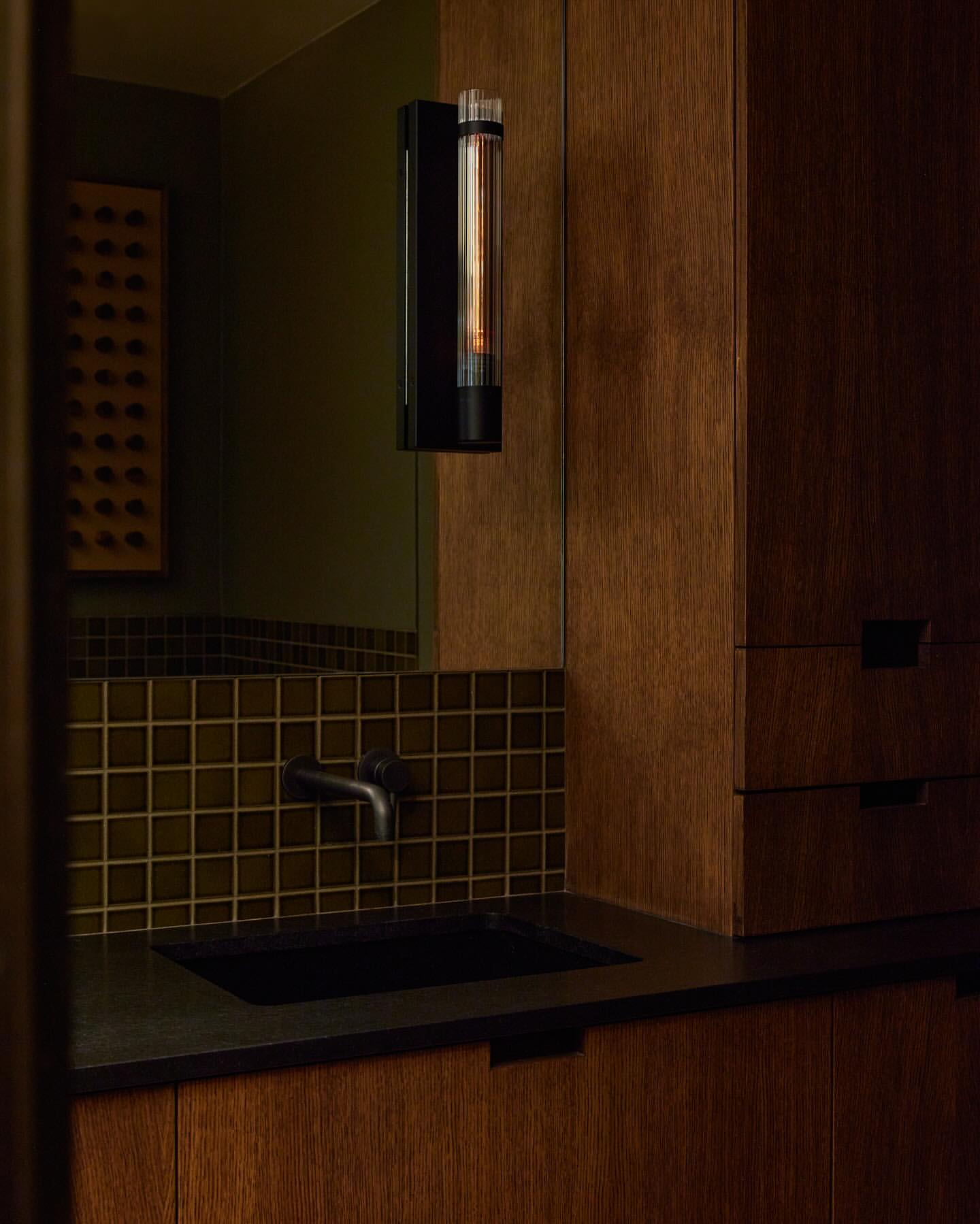 Modern bathroom interior featuring a dark sink, tiled backsplash, wall-mounted lighting fixture, and wood cabinets, creating a minimalist aesthetic.