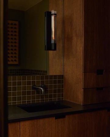 Modern bathroom interior featuring a dark sink, tiled backsplash, wall-mounted lighting fixture, and wood cabinets, creating a minimalist aesthetic.