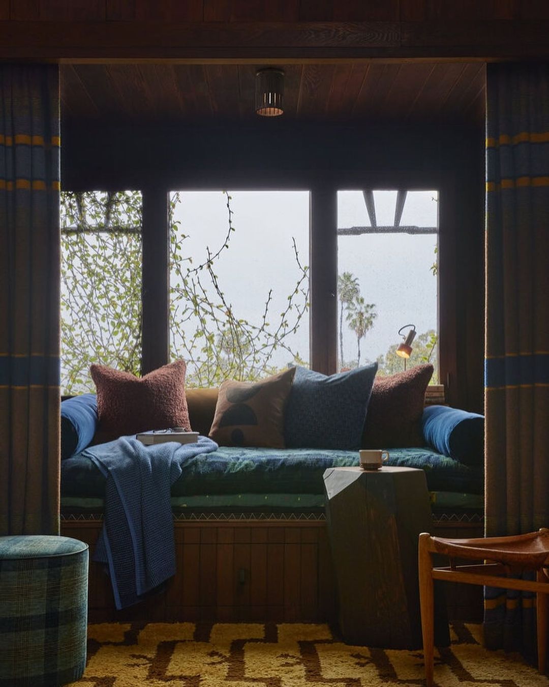 Daybed nook with pillows and blanket set inside a room with neutral tones and various textures, overlooking lush greenery through a window.