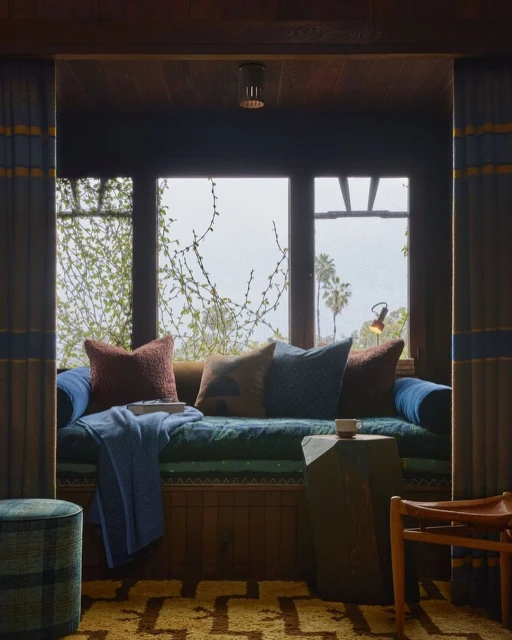 Daybed nook with pillows and blanket set inside a room with neutral tones and various textures, overlooking lush greenery through a window.
