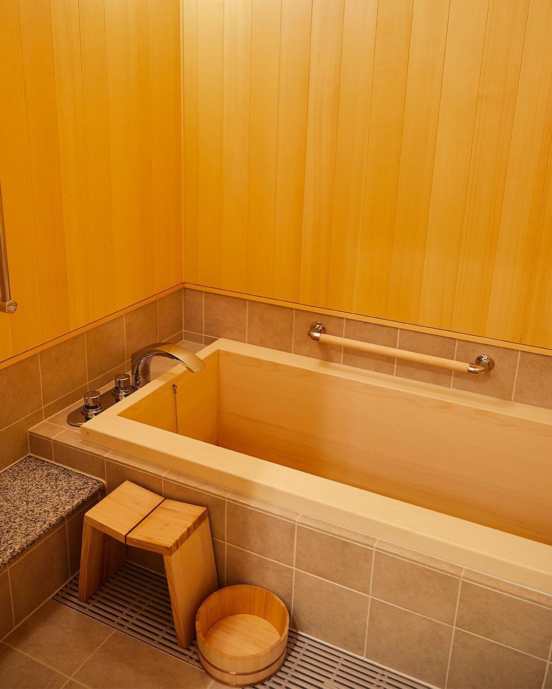 Indoor bathroom featuring a wooden bathtub, stool, and paneling, alongside tiled walls and flooring, creating a warm and minimalist aesthetic.