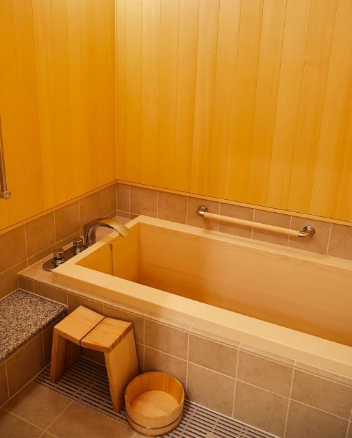 Indoor bathroom featuring a wooden bathtub, stool, and paneling, alongside tiled walls and flooring, creating a warm and minimalist aesthetic.