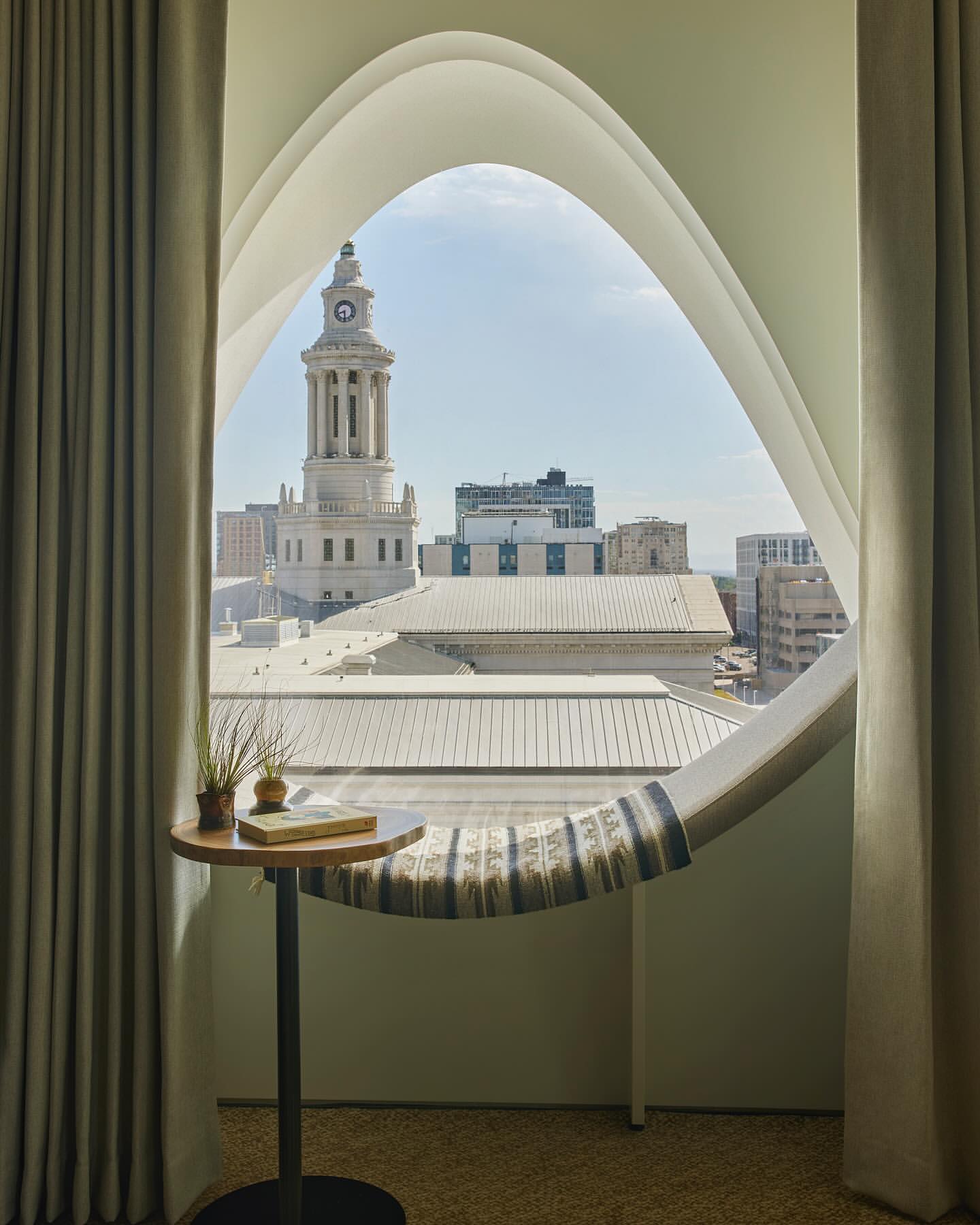 Interior view of a round wooden table next to a unique window overlooking a cityscape with a clock tower under a bright sky.