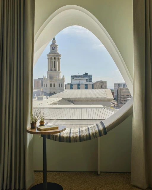 Interior view of a round wooden table next to a unique window overlooking a cityscape with a clock tower under a bright sky.