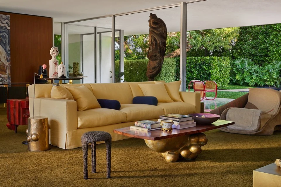 Living room interior featuring a light yellow sofa with bolster pillows, a unique coffee table, and view to a green outdoor area.