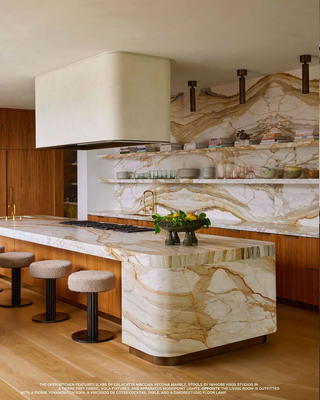 Modern kitchen interior featuring Calacatta Macchia Vecchia marble island, stools, and wood cabinets, illuminated by abundant lighting.