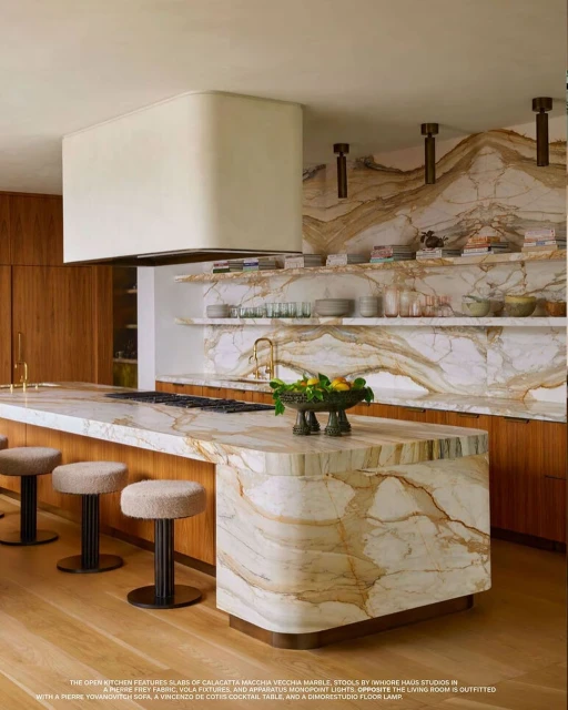 Modern kitchen interior featuring Calacatta Macchia Vecchia marble island, stools, and wood cabinets, illuminated by abundant lighting.