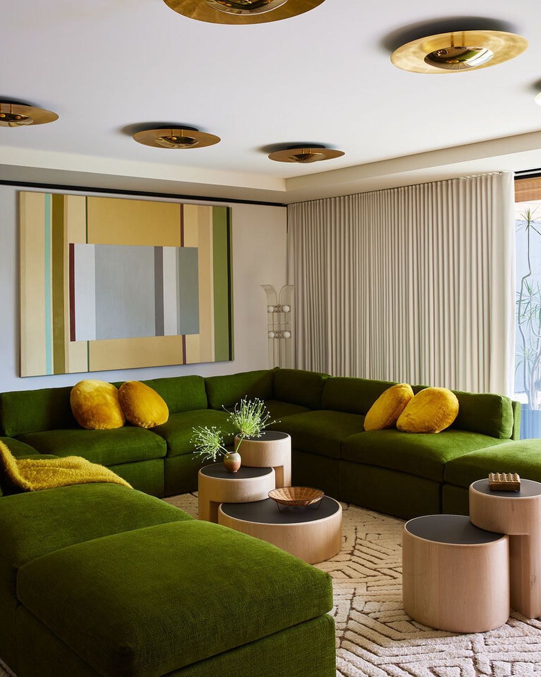 Living room interior featuring a large green sectional sofa with throw pillows, round coffee tables, patterned rug, and contemporary art.
