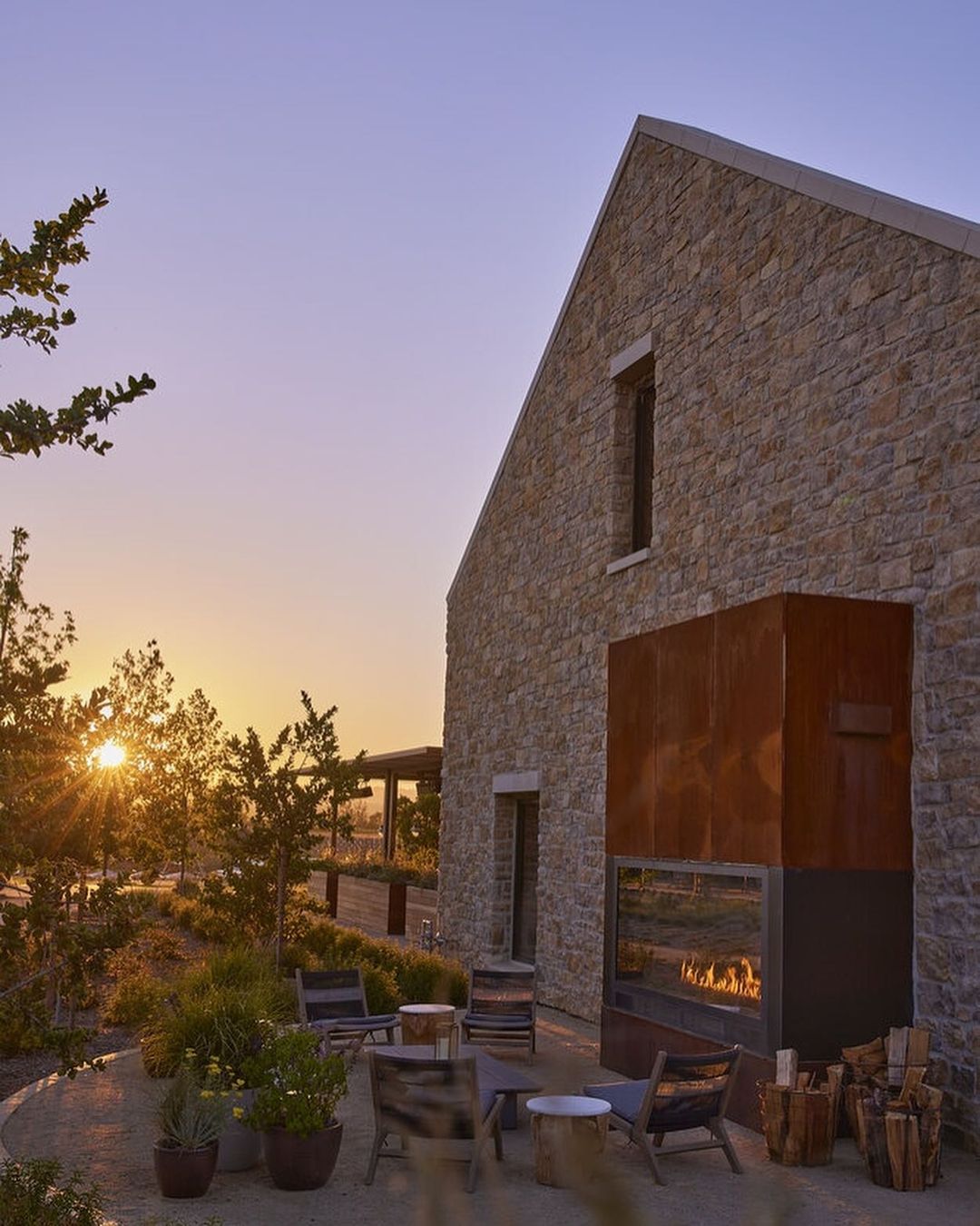 Exterior view of a stone building with a fireplace, surrounded by greenery and patio furniture at sunset.