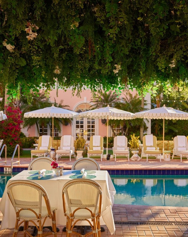 Outdoor dining table with wicker chairs and turquoise place settings next to a turquoise pool, parasols, and lounge chairs.