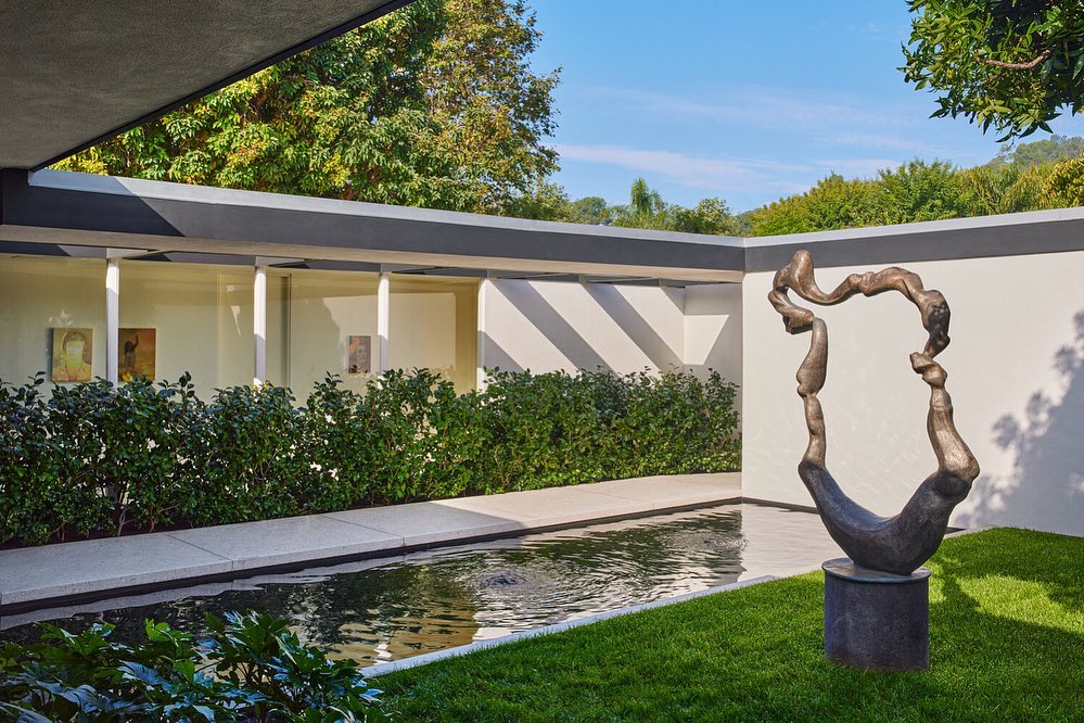 Exterior view of a modern building with a reflecting pool, an abstract sculpture, and green landscaping under a blue sky.