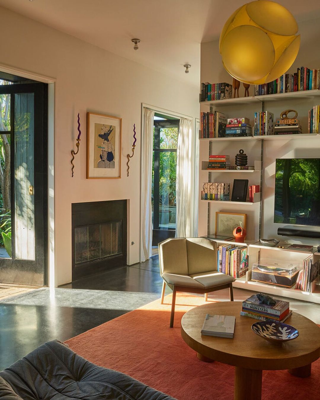 Living room interior with modern furniture, a round wooden coffee table on a red rug, a beige chair, and a bookshelf filled with books.