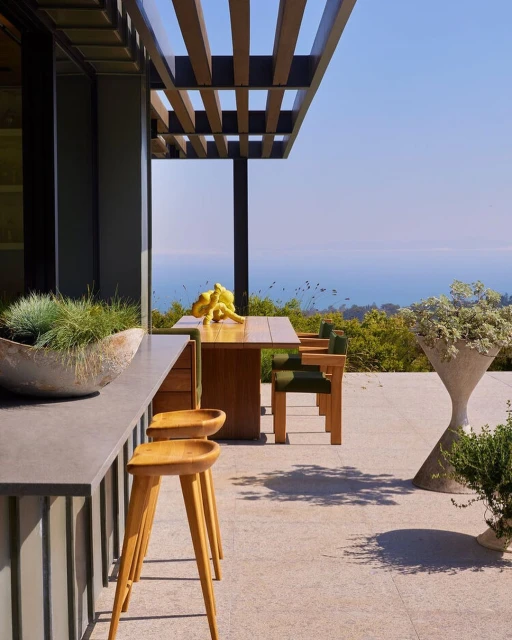 Outdoor patio with wooden table, chairs, and stools, featuring modern design, plants, and a view of the ocean under a clear sky.