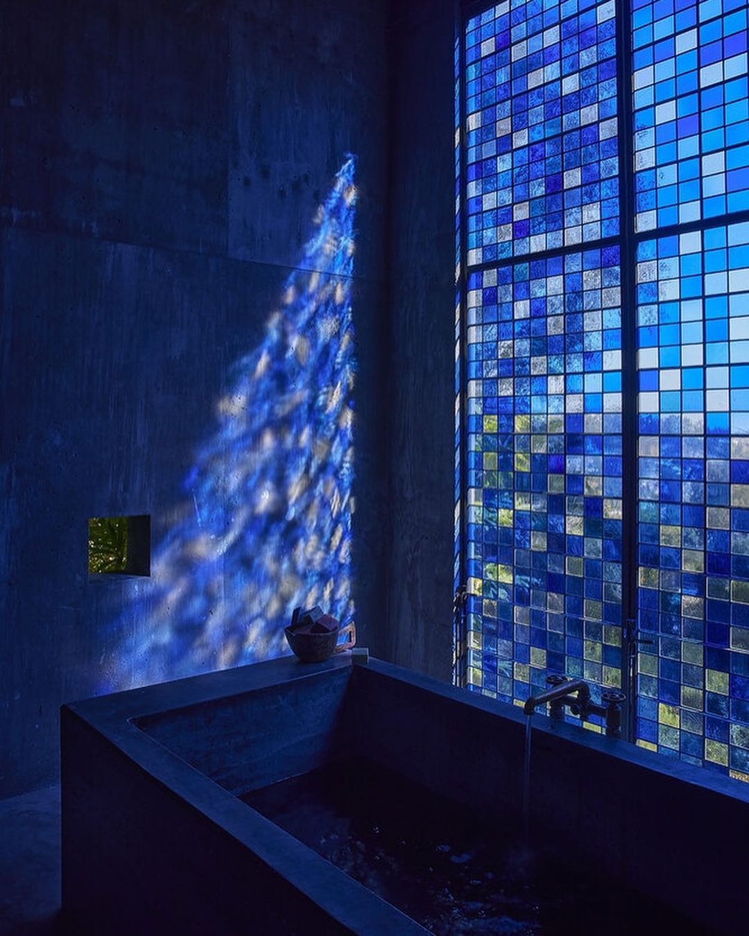 Modern bathroom interior with a concrete bathtub, square blue-toned window, and dappled sunlight pattern on the wall, creating a serene and minimalist space.