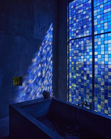 Modern bathroom interior with a concrete bathtub, square blue-toned window, and dappled sunlight pattern on the wall, creating a serene and minimalist space.