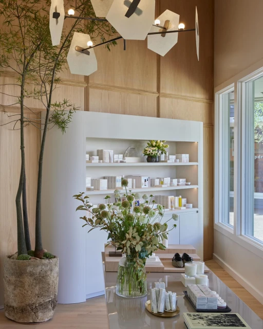 A retail display area with skincare products and a flower arrangement on a table in an indoor setting featuring neutral wall tones and natural light.