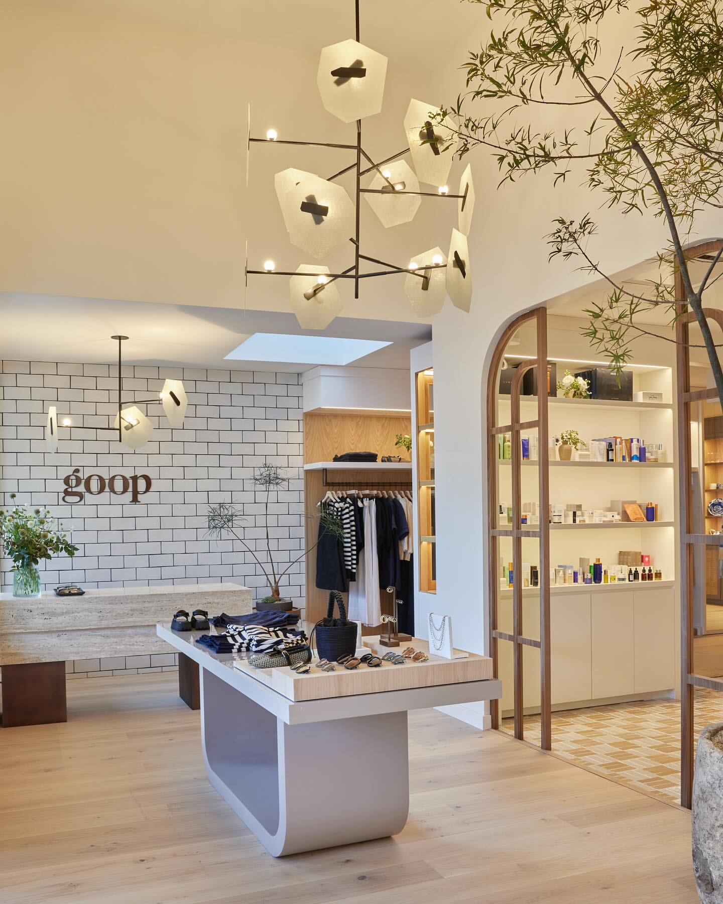 Interior of a Goop store featuring clothing, beauty products, and home goods displayed on tables and shelves with unique lighting fixtures.
