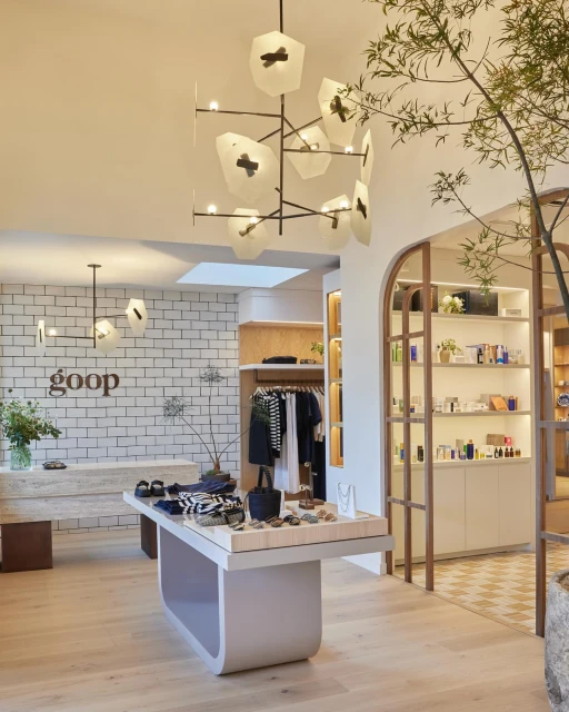 Interior of a Goop store featuring clothing, beauty products, and home goods displayed on tables and shelves with unique lighting fixtures.