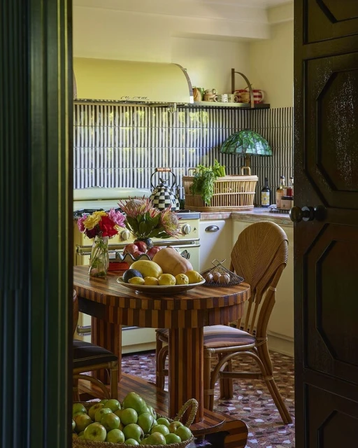 Still life view of a kitchen with a wooden table and rattan chair, featuring a fruit bowl, flowers, patterned tiles, and vintage stove.