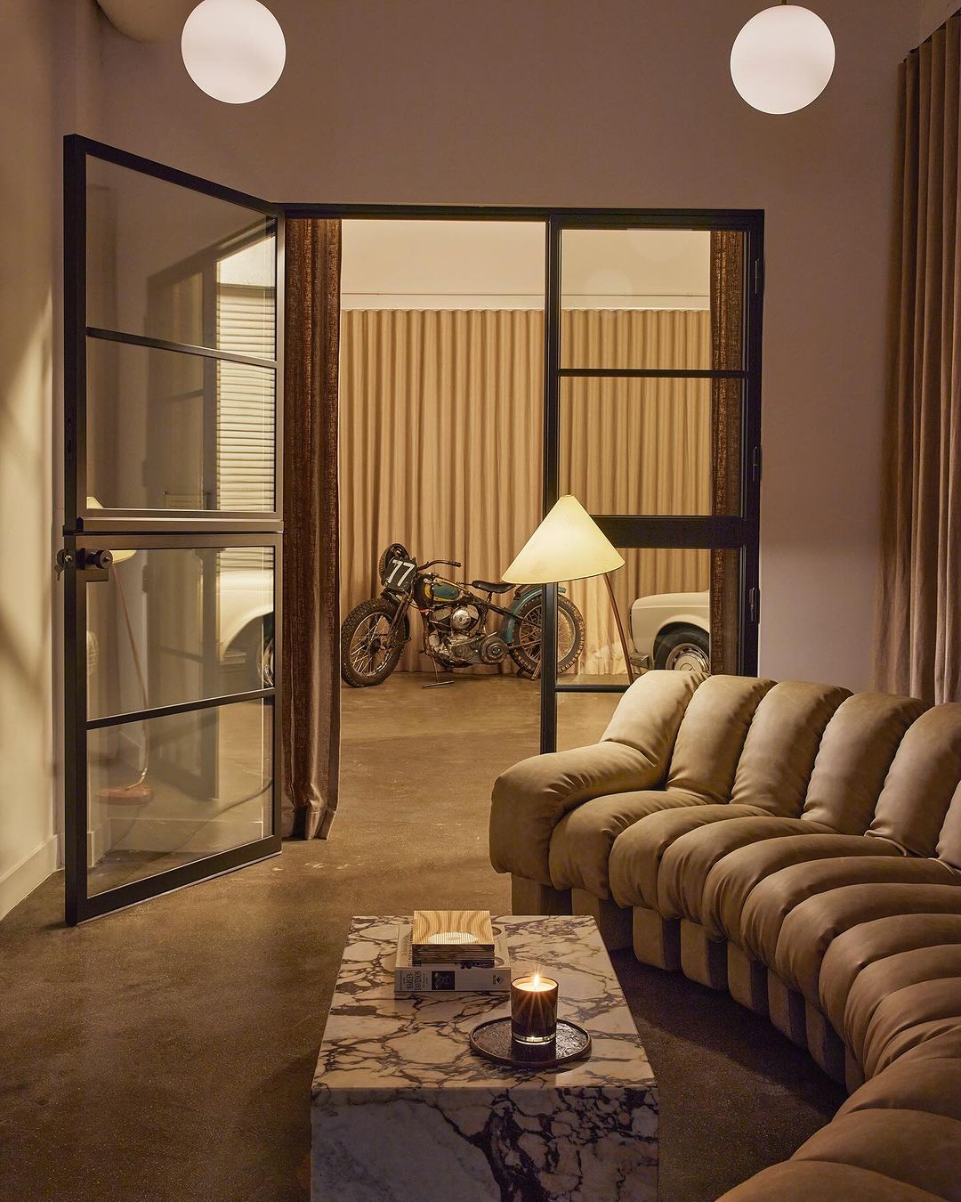 Tan sectional sofa and marble coffee table in an indoor living room scene with vintage motorcycle and cars visible through black frame doors.