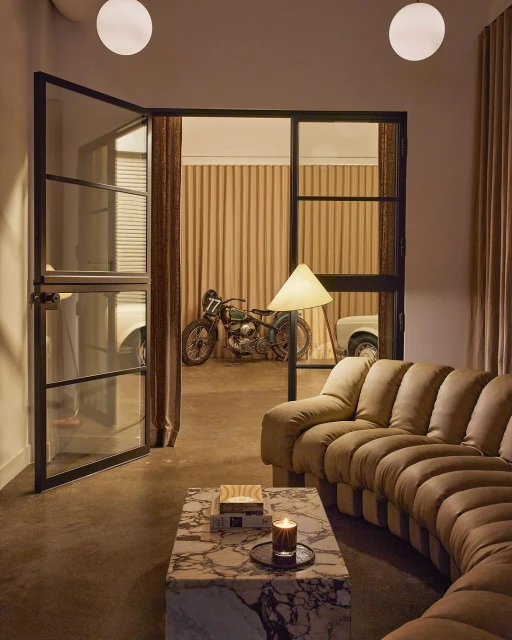 Tan sectional sofa and marble coffee table in an indoor living room scene with vintage motorcycle and cars visible through black frame doors.