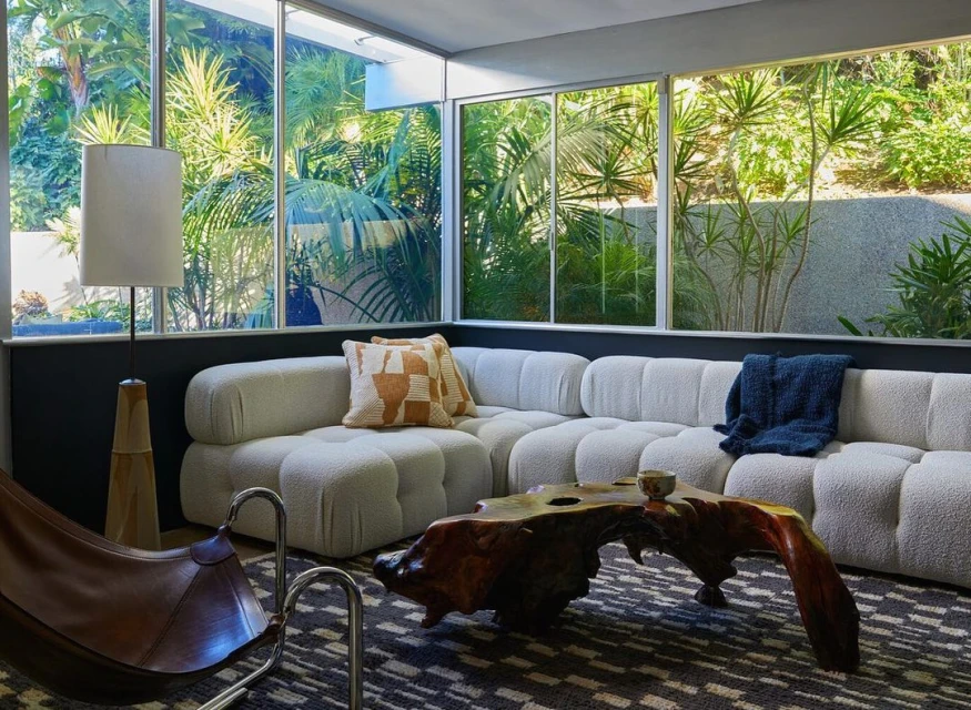 Living room interior with a modern white sofa, wooden coffee table, floor lamp, and a large window overlooking lush greenery.