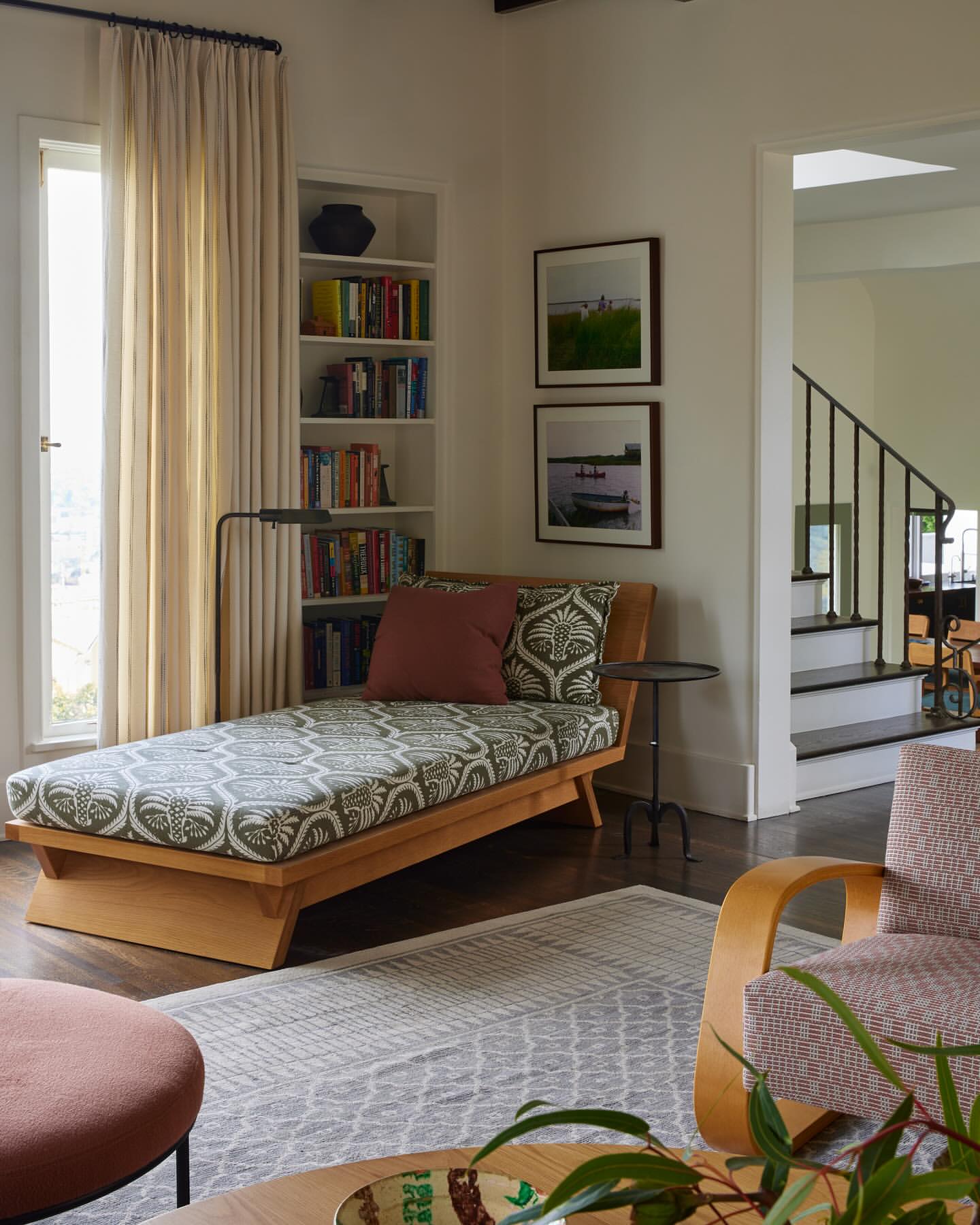 Interior view of a living room with a wooden chaise lounge featuring patterned fabric, flanked by a bookcase and a stairway.