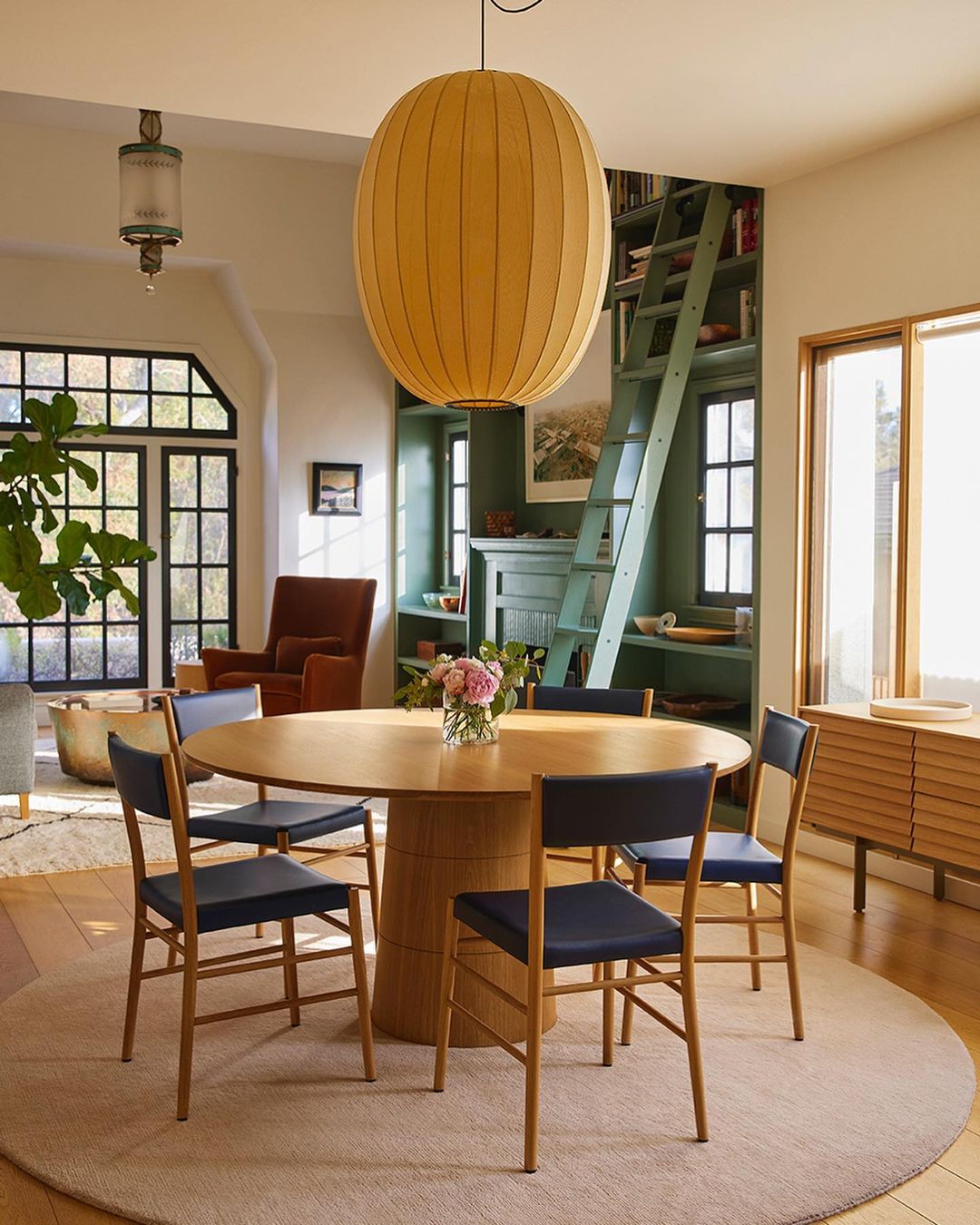 Interior of a dining room featuring a round wooden table with blue chairs, a large paper lantern pendant, and a built-in bookcase.