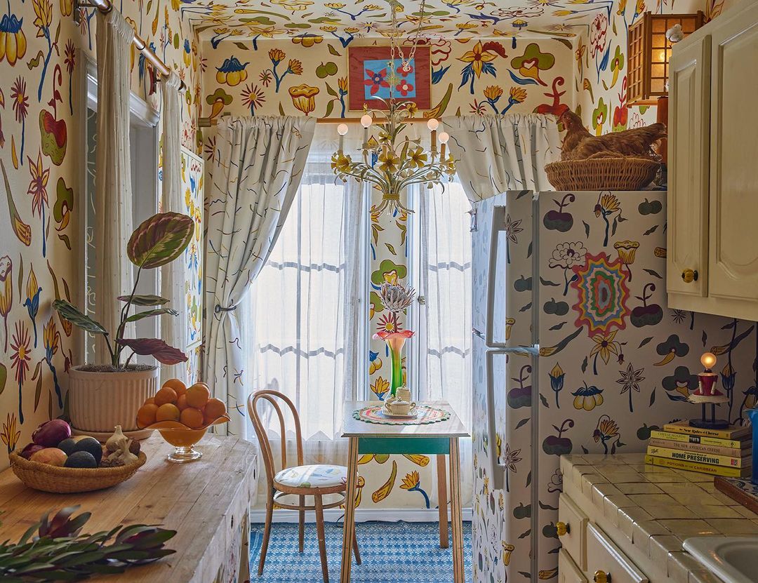 Interior shot of a brightly decorated kitchen with floral wallpaper, patterned floors, and a wooden table with fruit and flowers.