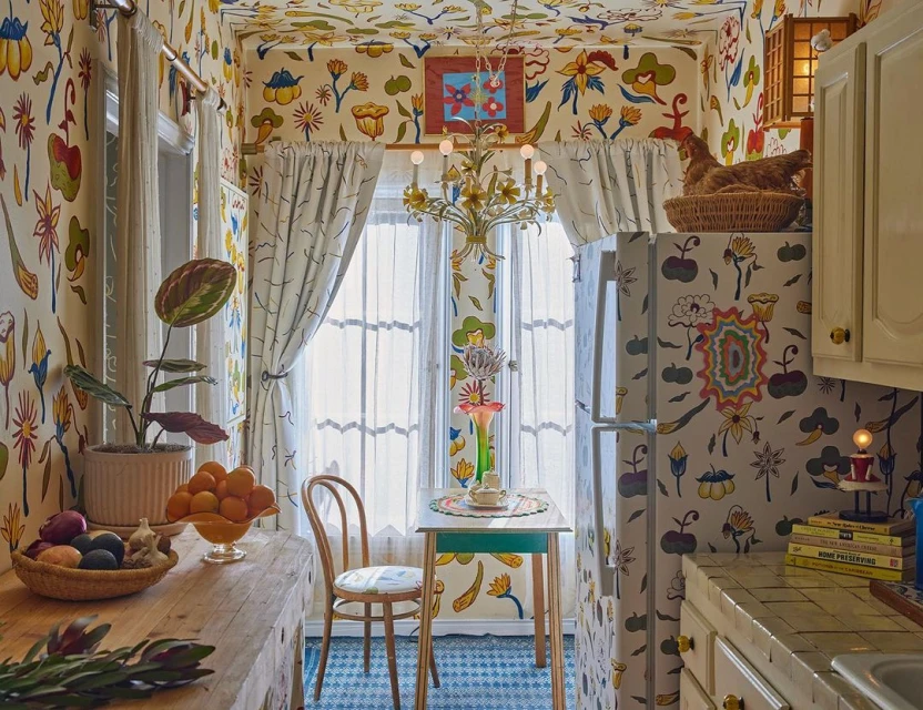 Interior shot of a brightly decorated kitchen with floral wallpaper, patterned floors, and a wooden table with fruit and flowers.
