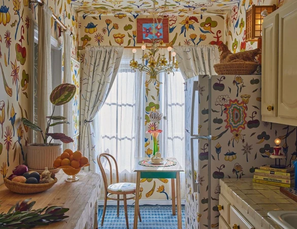 Interior shot of a brightly decorated kitchen with floral wallpaper, patterned floors, and a wooden table with fruit and flowers.