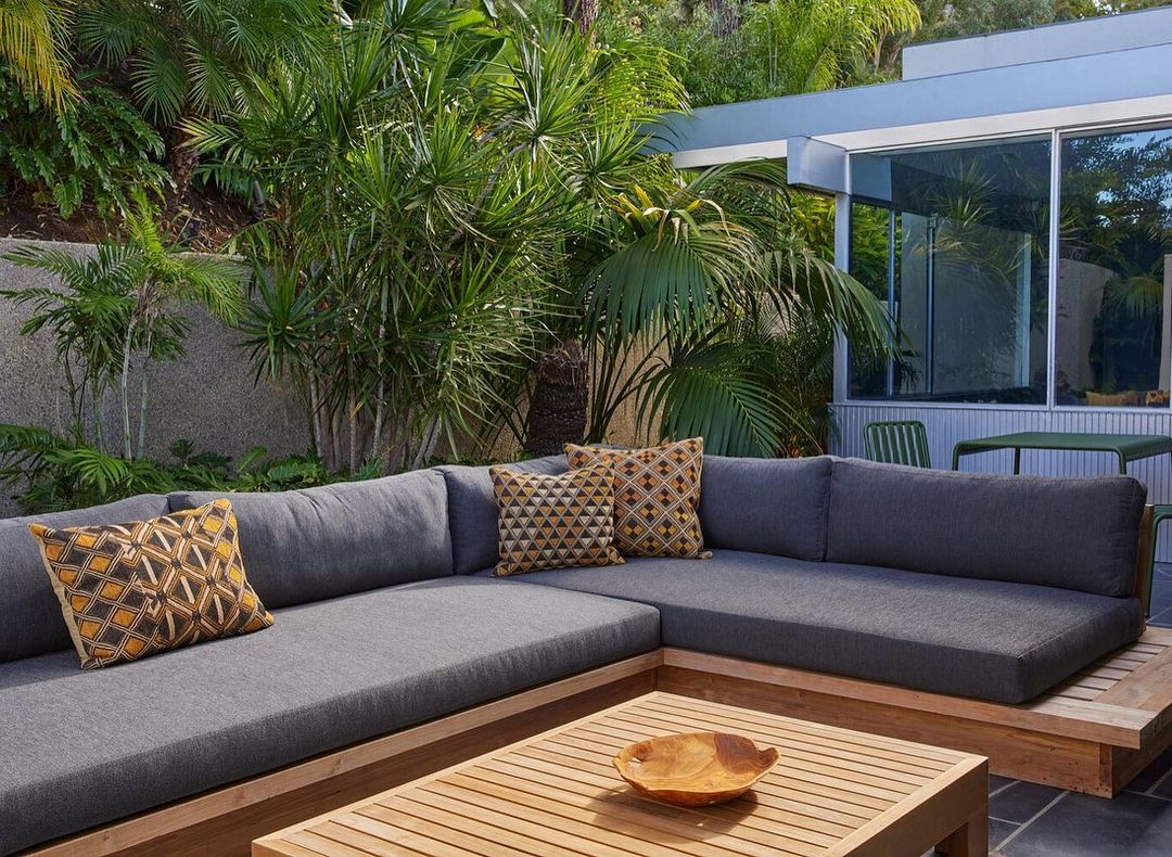 Outdoor living space featuring a grey sectional sofa with patterned cushions, wooden coffee table, and lush tropical plants.