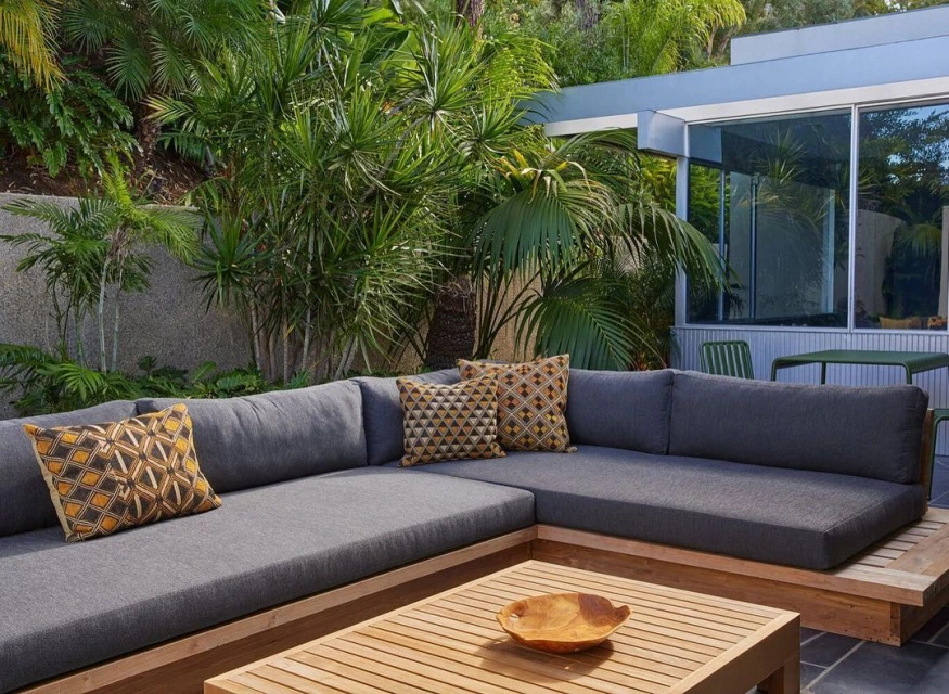 Outdoor living space featuring a grey sectional sofa with patterned cushions, wooden coffee table, and lush tropical plants.