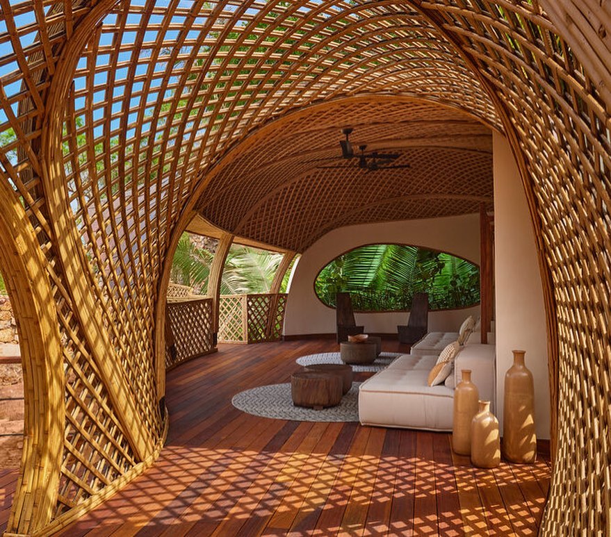 Indoor lounge area featuring a bamboo-constructed archway and ceiling, hardwood floors, white cushions, cylindrical stools, and ambient lighting.