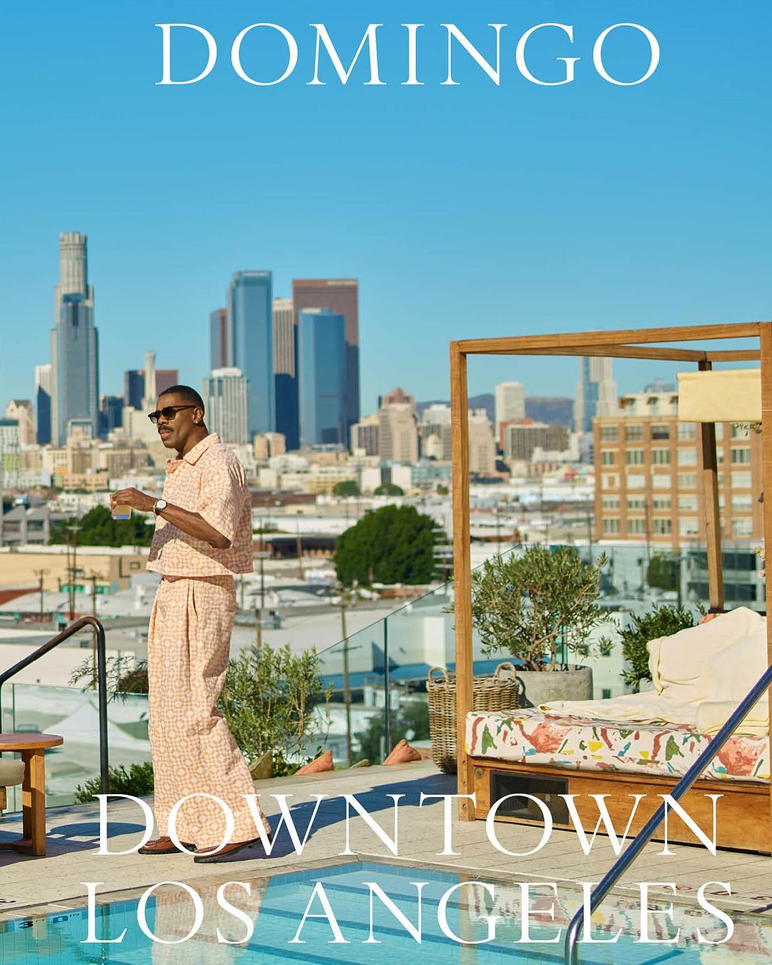 Man in patterned peach suit stands on rooftop with drink, with Downtown Los Angeles skyline and swimming pool in bright daytime shot.
