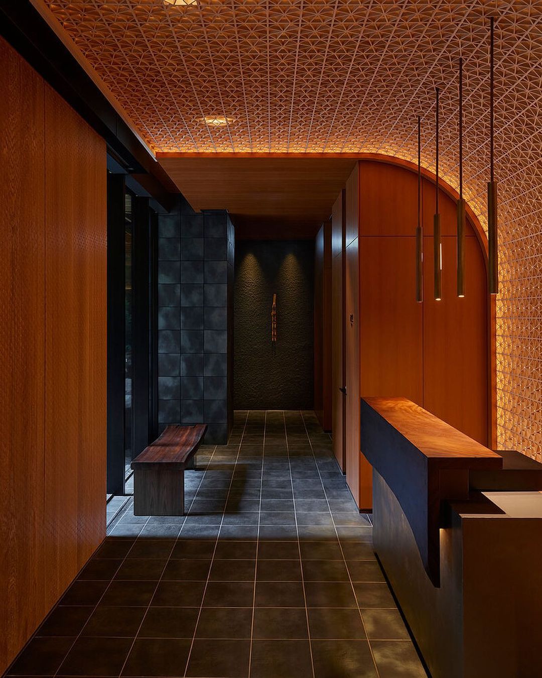 Interior view of a modern hallway with tiled floors, wood paneling, a textured ceiling, and a reception desk in warm, ambient lighting.