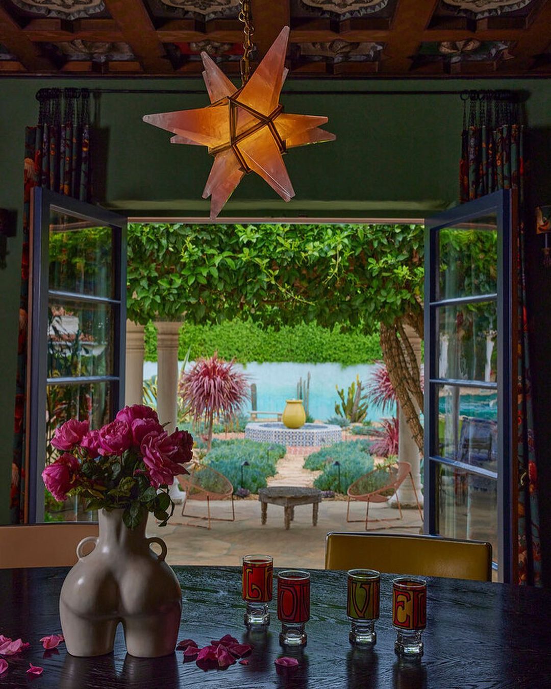 Indoor dining room features pink roses in a white vase on a dark table, with colorful shot glasses and an open window overlooking a garden.