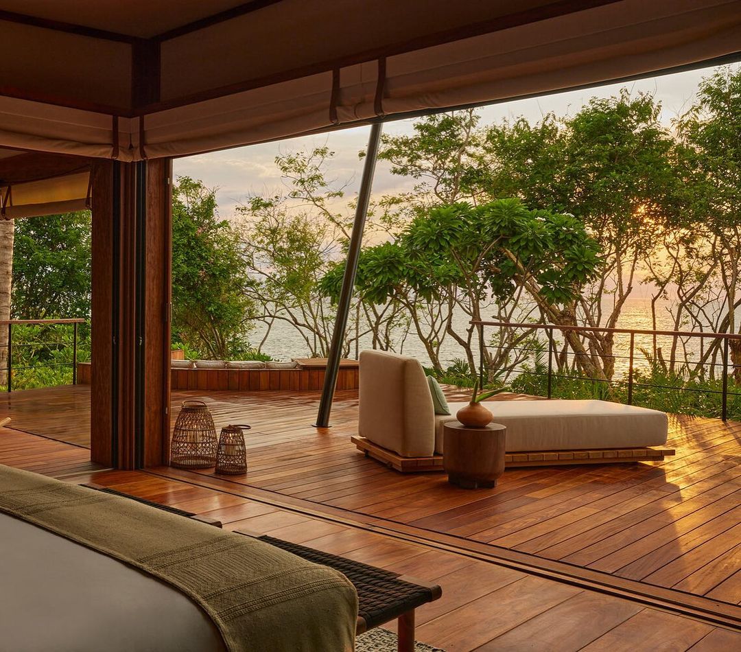 Relaxing lounge area on wooden deck with ocean view through trees. Neutral furniture, warm lighting, indoor-outdoor living, peaceful scene.