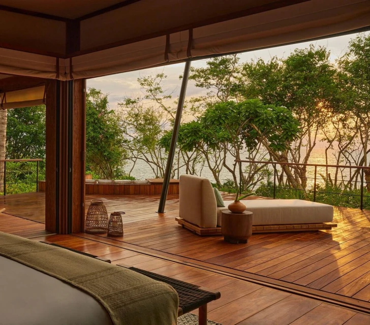 Relaxing lounge area on wooden deck with ocean view through trees. Neutral furniture, warm lighting, indoor-outdoor living, peaceful scene.
