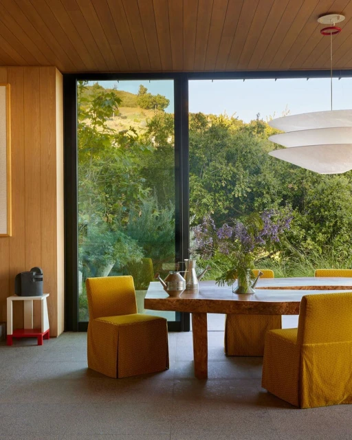 Dining room interior featuring a natural wood table with yellow upholstered chairs, a modern chandelier, and a large window view.