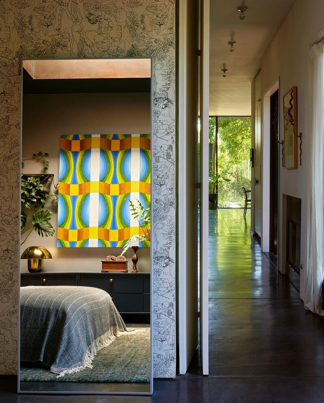 Bedroom interior reflected in mirror: bed with grey cover, geometric art, designer lamp, wallpaper in interior hallway of house.