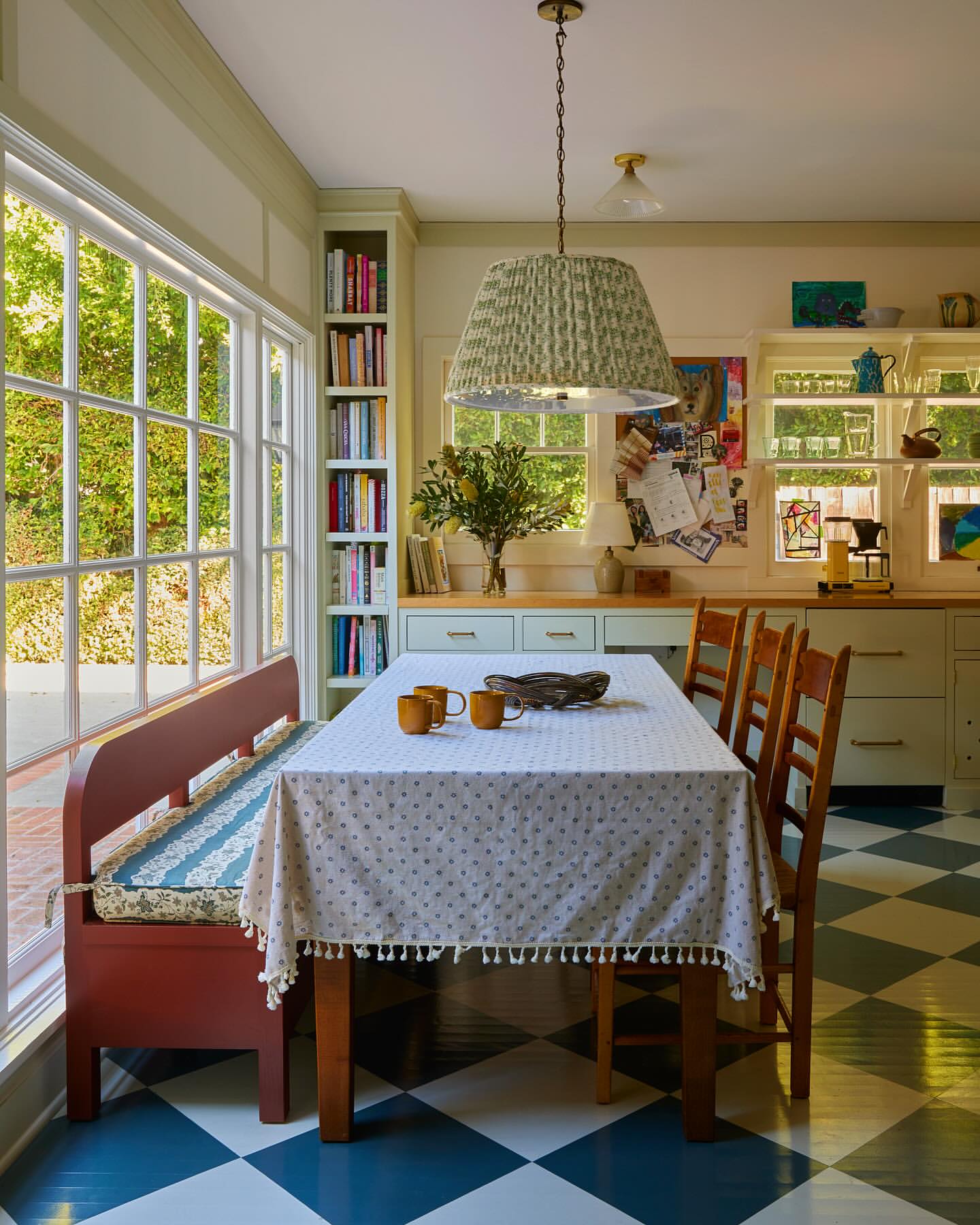 Cozy kitchen interior featuring a dining table with a floral tablecloth, surrounded by wooden chairs and a bench by a large window.