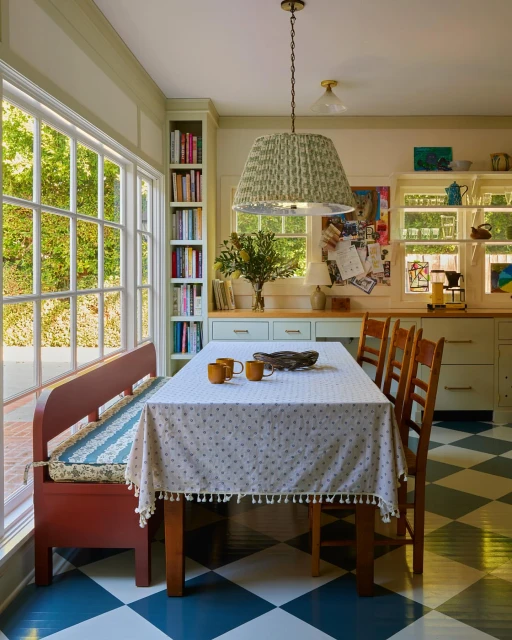 Cozy kitchen interior featuring a dining table with a floral tablecloth, surrounded by wooden chairs and a bench by a large window.