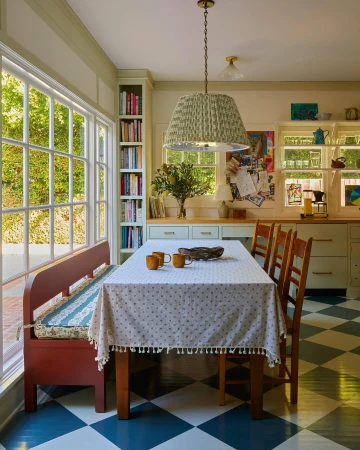 Cozy kitchen interior featuring a dining table with a floral tablecloth, surrounded by wooden chairs and a bench by a large window.