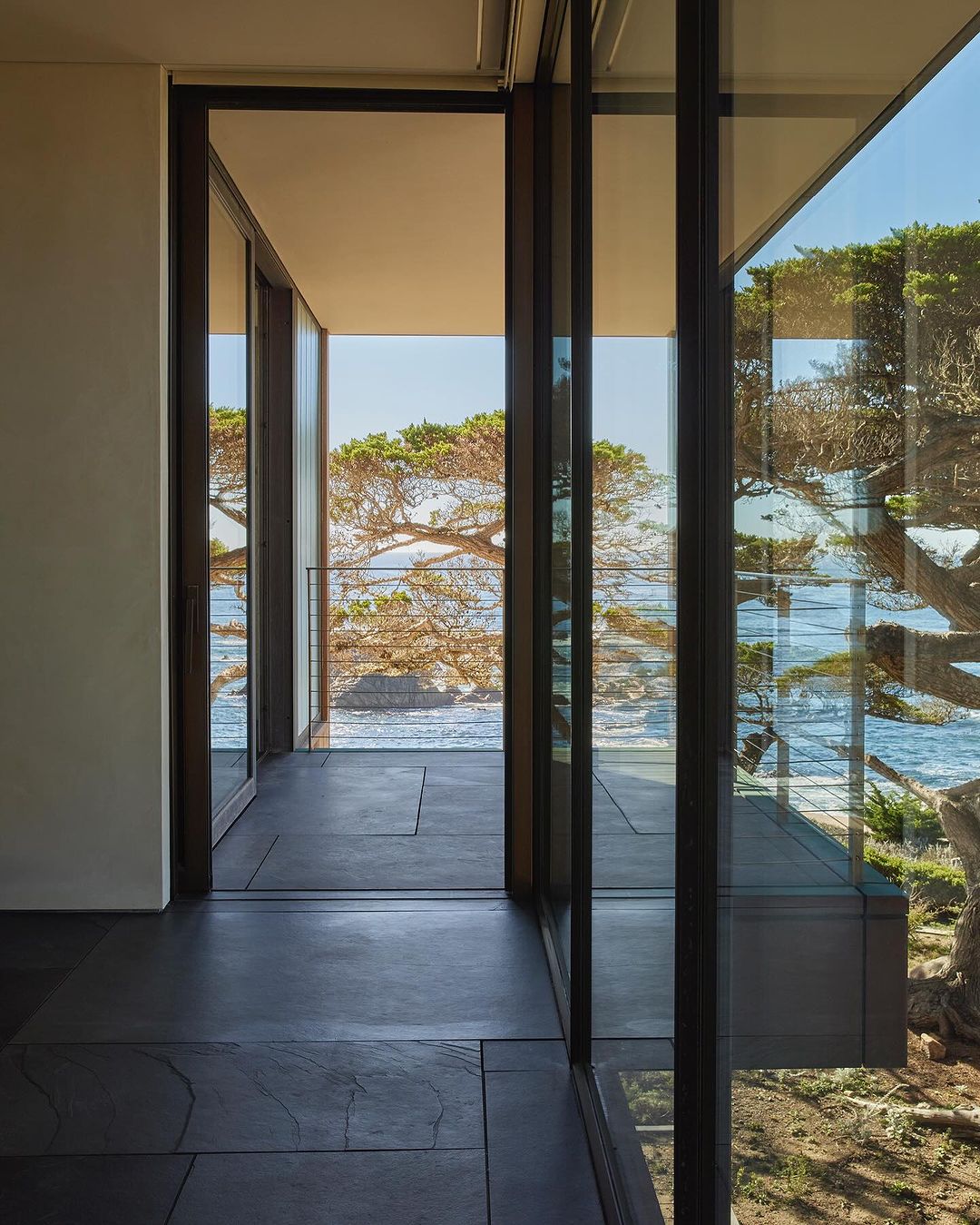 Modern home interior showcases floor-to-ceiling windows with ocean view, and tree. Dark tiled floor leads to an outdoor balcony.