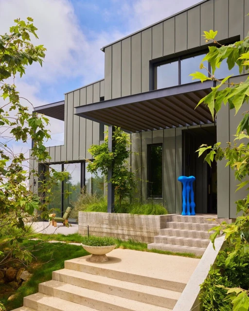 Exterior view of a modern two-story house with gray vertical siding, concrete stairs, and an abstract sculpture on the porch.