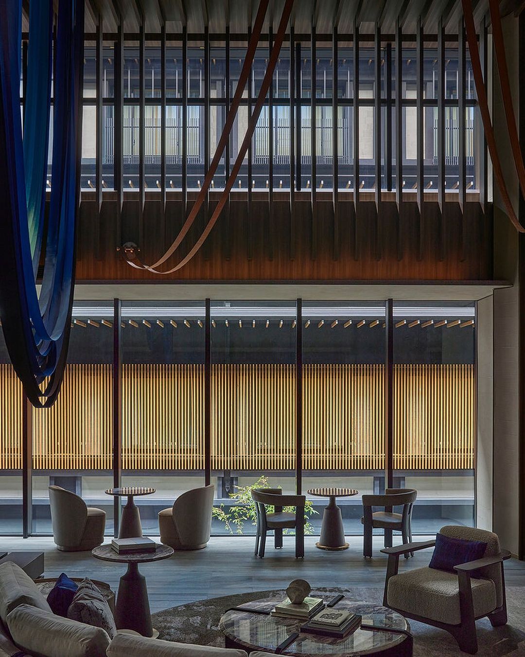 Interior view of a modern lounge with armchairs and tables arranged on a rug in front of a window with a wood slat screen.